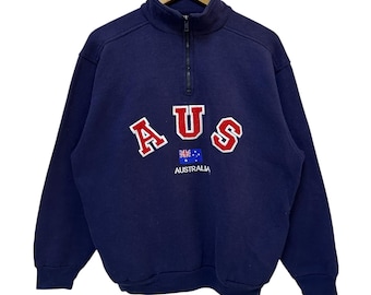 May include: Navy blue sweatshirt with a quarter-zip collar. The front features the word "AUS" in red block letters, with the Australian flag and the word "AUSTRALIA" underneath. The sweatshirt is hanging on a wooden hanger.