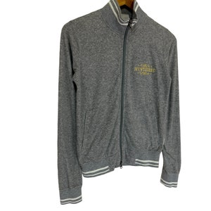 May include: A gray zip-up jacket with white ribbed trim on the collar, cuffs, and hem. The jacket has a gold embroidered logo that says "HYSTERIC" on the front.