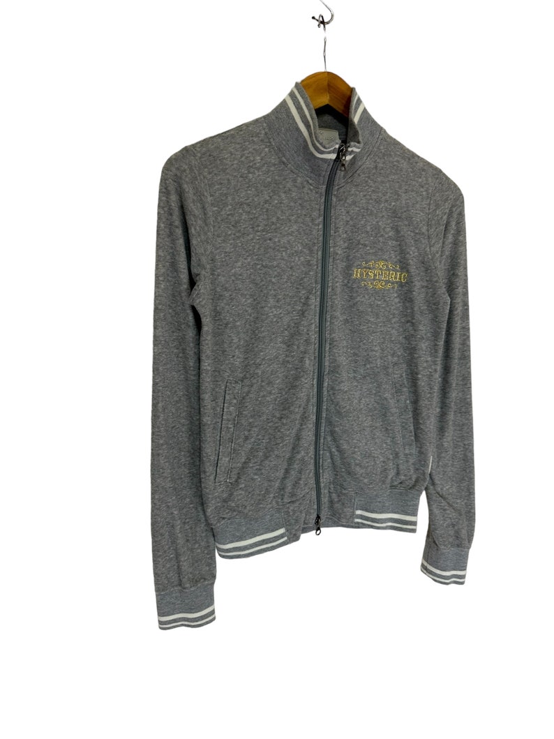 May include: A gray zip-up jacket with white ribbed trim on the collar, cuffs, and hem. The jacket has a gold embroidered logo that says "HYSTERIC" on the front.