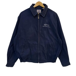 May include: Navy blue zip-up jacket with a collar and two front pockets. The jacket features the UD Trucks logo and the text "NISSAN DIESEL AMERICA" in white on the chest. The jacket is hanging on a wooden hanger.