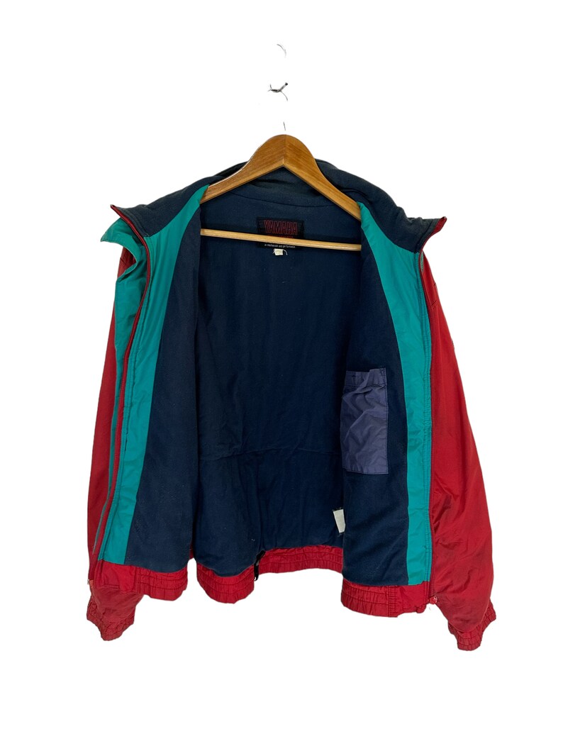 May include: A red, teal and navy blue jacket with a zip closure. The jacket has a small pocket on the inside. The label on the jacket reads "Warm Up".