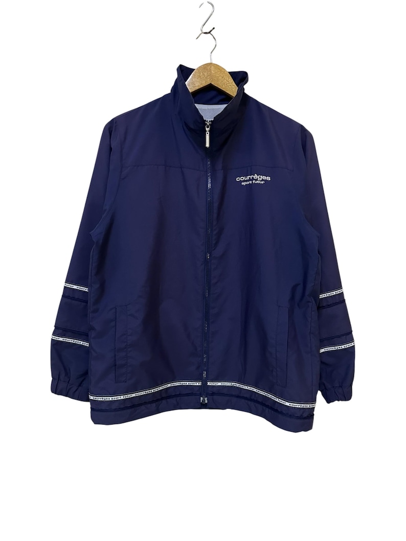 May include: Navy blue Courr&egrave;ges sport futur jacket with a full-length zipper and a stand-up collar. The jacket features white stripes with text on the sleeves and bottom hem. The brand name is printed in white on the chest.