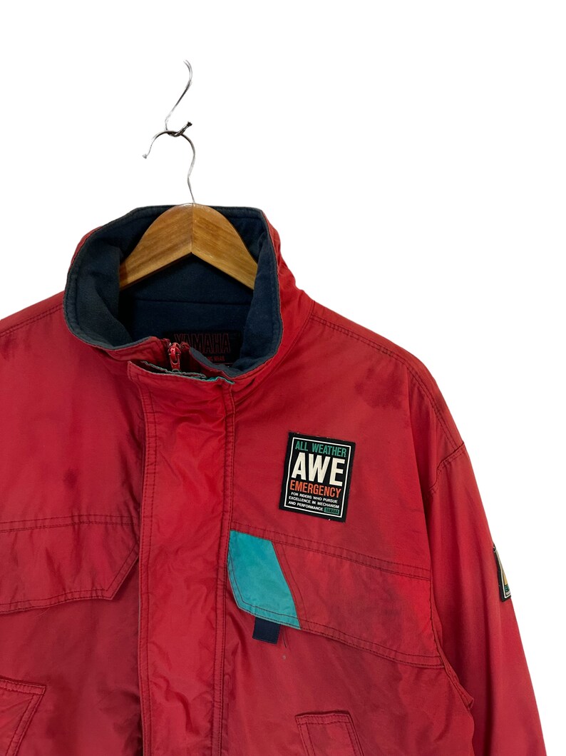 May include: Red jacket with a blue and teal pocket and a patch that says "All Weather AWE Emergency" with a logo underneath.