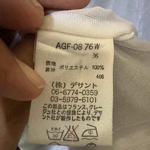 May include: A white clothing tag with black text, held by a hand. The tag includes the text "AGF-08 76 W", "36", and Japanese characters. It also lists the material as 100% polyester and includes washing instructions.