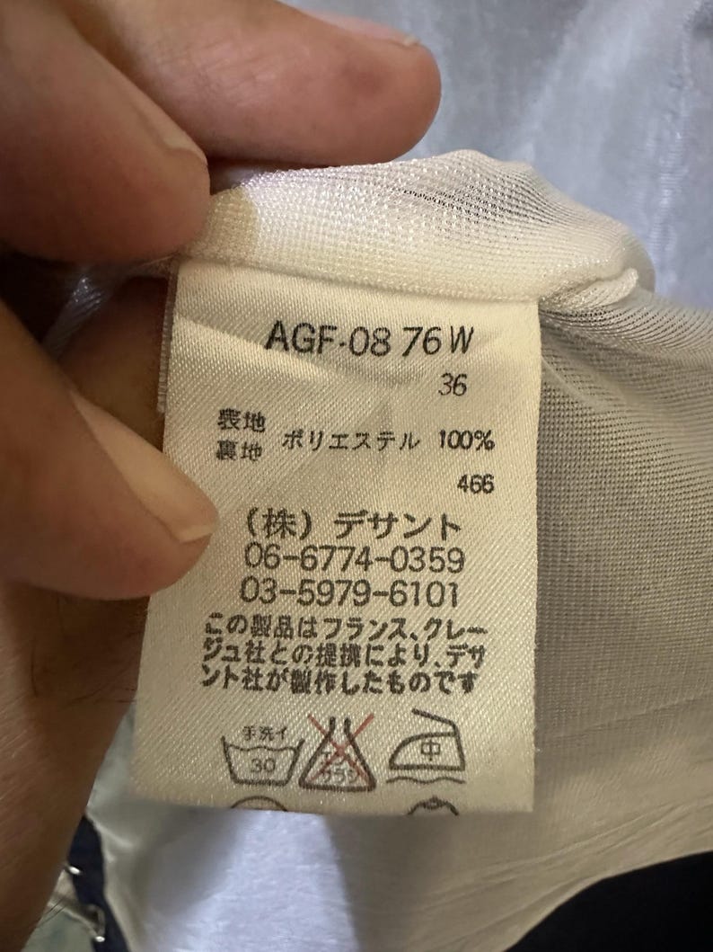 May include: A white clothing tag with black text, held by a hand. The tag includes the text "AGF-08 76 W", "36", and Japanese characters. It also lists the material as 100% polyester and includes washing instructions.