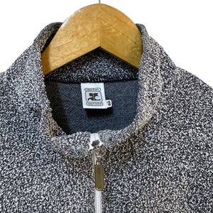 May include: A gray and white speckled zip-up jacket with a high collar. The jacket features a silver zipper and a small label with the text "courr&egrave;ges sport futur". The jacket is hanging on a wooden hanger.