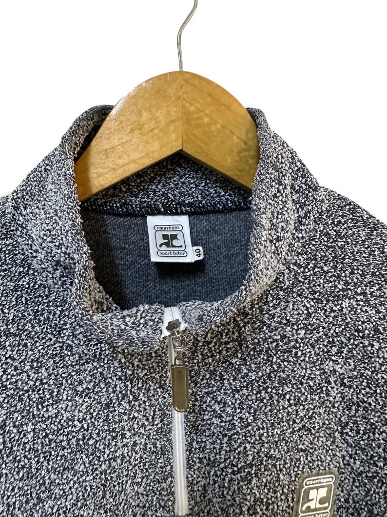 May include: A gray and white speckled zip-up jacket with a high collar. The jacket features a silver zipper and a small label with the text "courr&egrave;ges sport futur". The jacket is hanging on a wooden hanger.