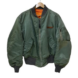 Puede incluir: Chaqueta bomber verde oliva con cuello, pu&ntilde;os y cintura gris oscuro. La chaqueta tiene cierre de cremallera, dos bolsillos delanteros y un peque&ntilde;o parche en el pecho. El forro interior es de color naranja brillante.