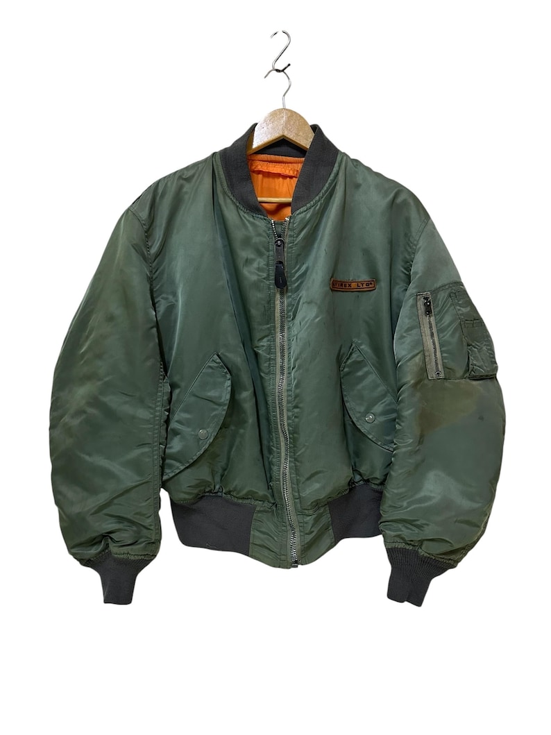 Puede incluir: Chaqueta bomber verde oliva con cuello, pu&ntilde;os y cintura gris oscuro. La chaqueta tiene cierre de cremallera, dos bolsillos delanteros y un peque&ntilde;o parche en el pecho. El forro interior es de color naranja brillante.