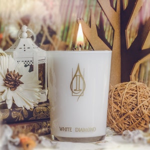 May include: White candle with a gold label that reads "White Diamond" and a gold logo of a stylized water drop. The candle is lit and the flame is visible. The candle is on a table with other decorative items.