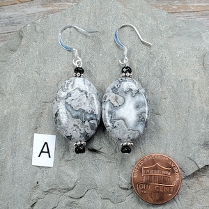 May include: A pair of dangle earrings featuring oval-shaped gray and white patterned stones. Each earring is accented with small black beads and silver-toned metal findings, suspended from silver-toned ear wires. A small card with the letter A is visible.