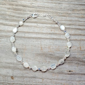 May include: A delicate bracelet featuring oval, off-white beads interspersed with small, clear and blue crystals. The bracelet has a silver-colored chain and clasp. The beads are smooth and have a subtle sheen, creating an elegant accessory.