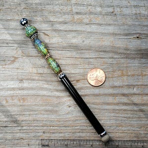May include: A decorative stylus with a black barrel and a beaded top. The top features green, blue, and gold beads with a silver accent. A penny is next to the stylus for size reference. The stylus is approximately 7 inches long.