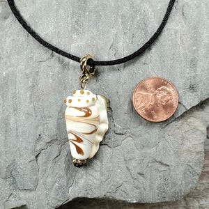 May include: A seashell pendant necklace with a black cord. The pendant is white and brown with gold accents. A penny is next to the pendant for size reference. The necklace is on a gray stone surface.