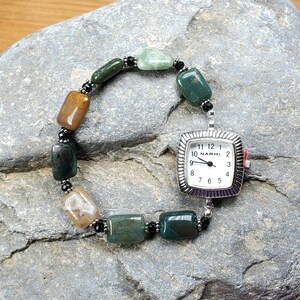 May include: A silver-toned watch with a square face and a white dial. The watch is attached to a bracelet made of green, brown, and beige rectangular beads. The beads are strung together with black beads. The watch face reads "NARMI".