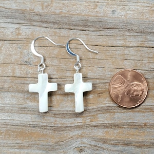 May include: A pair of white cross earrings with silver hooks. The earrings are displayed on a wooden surface next to a penny. The crosses have a pearlescent sheen.