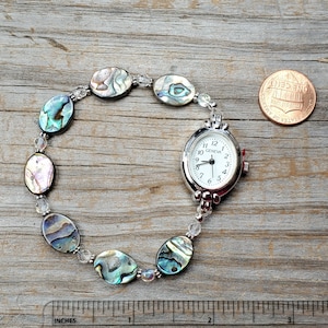 May include: A silver-toned Geneva watch with a white face and black numerals, connected to a bracelet of iridescent abalone shell beads and clear crystal beads. The watch is next to a penny and a ruler showing inches.