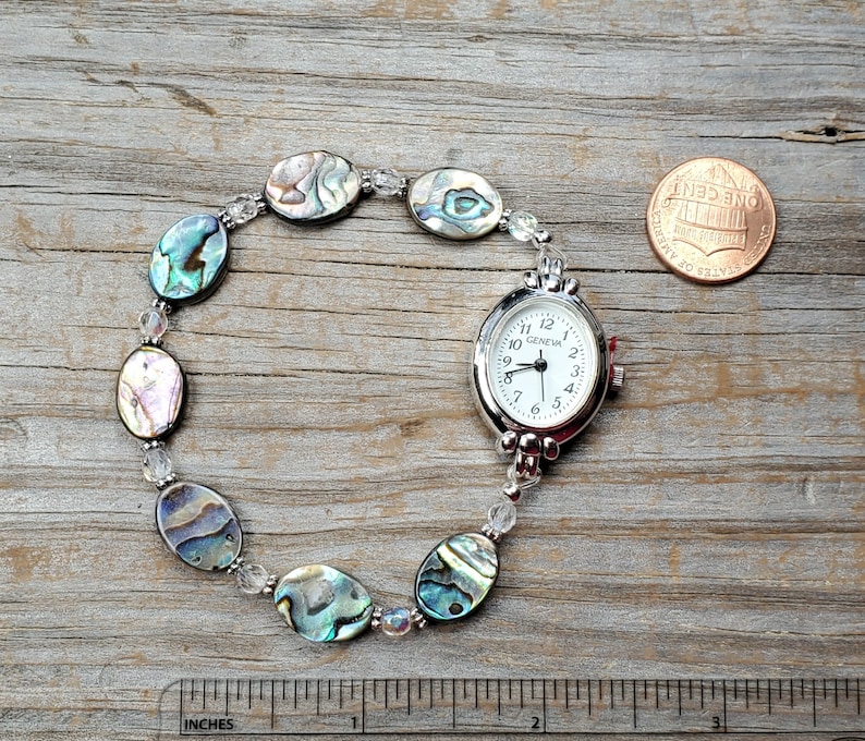 May include: A silver-toned Geneva watch with a white face and black numerals, connected to a bracelet of iridescent abalone shell beads and clear crystal beads. The watch is next to a penny and a ruler showing inches.