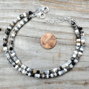 May include: Two beaded bracelets with a mix of black, white, and gray beads, along with a few black beads. The bracelets are connected by a silver chain and clasps. A penny is placed near the bracelets for size comparison. The bracelets are on a weathered wooden surface.