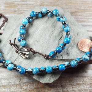 May include: A handmade necklace and bracelet set featuring vibrant blue gemstone beads. The necklace and bracelet are strung on brown leather cord, with silver-toned metal accents. A small seashell charm and a coin are also visible.