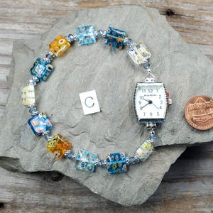 May include: A watch bracelet featuring a silver-tone Narmi watch face with a white dial and black numerals, connected to a series of square, multi-colored millefiori glass beads and small silver-tone spacer beads, positioned next to a copper penny.
