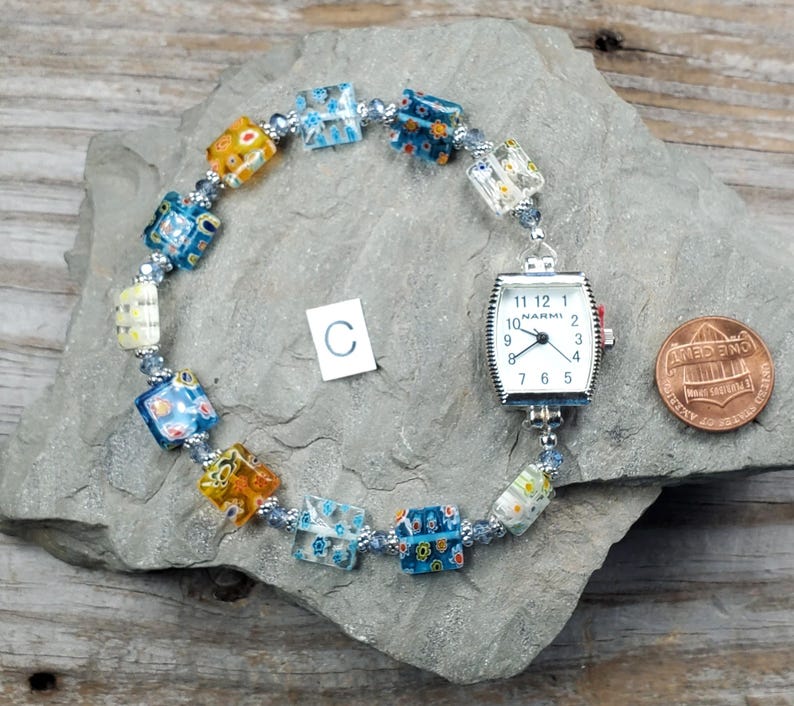 May include: A watch bracelet featuring a silver-tone Narmi watch face with a white dial and black numerals, connected to a series of square, multi-colored millefiori glass beads and small silver-tone spacer beads, positioned next to a copper penny.