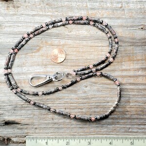 Pode incluir: Um cord&atilde;o de contas com um fecho prateado. O cord&atilde;o apresenta contas cil&iacute;ndricas cinzentas e pretas intercaladas com pequenas contas cor-de-rosa. Uma moeda est&aacute; ao lado para compara&ccedil;&atilde;o de tamanho. O cord&atilde;o est&aacute; sobre uma superf&iacute;cie de madeira.