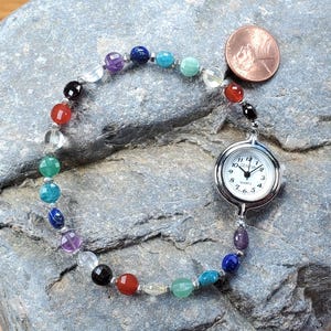 May include: A wristwatch featuring a round silver-tone case with a white analog face labeled Geneva Quartz, connected to a beaded bracelet made of multi-colored gemstone spheres in shades of red, blue, green, purple, and clear, next to a penny.