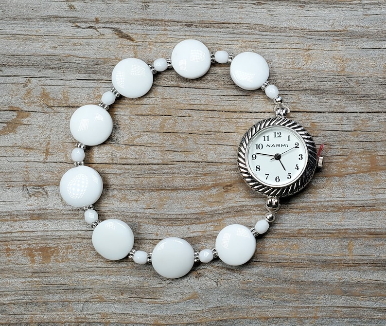 Puede incluir: Pulsera de cuentas blancas con un reloj plateado adjunto. La esfera del reloj es blanca con agujas negras y n&uacute;meros negros. La esfera del reloj dice "NARMI" en la parte superior.