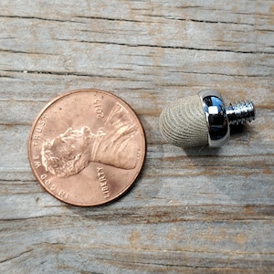 May include: A small, silver-coloured, threaded, round, fabric-covered object with a shiny metal top. The object is about the size of a UK penny.