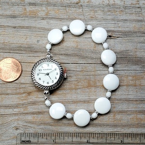 Puede incluir: Un reloj de pulsera con cuentas blancas y una esfera plateada que dice "NARMI".