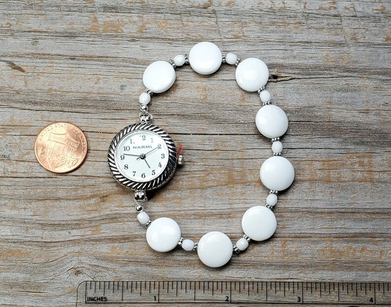 Puede incluir: Un reloj de pulsera con cuentas blancas y una esfera plateada que dice "NARMI".