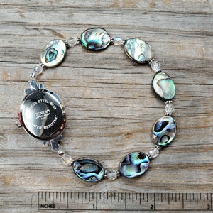 May include: A Geneva watch with a stainless steel back and water-resistant feature. The watch is attached to a bracelet made of iridescent abalone shells and clear beads. The bracelet is approximately 7 inches in length.