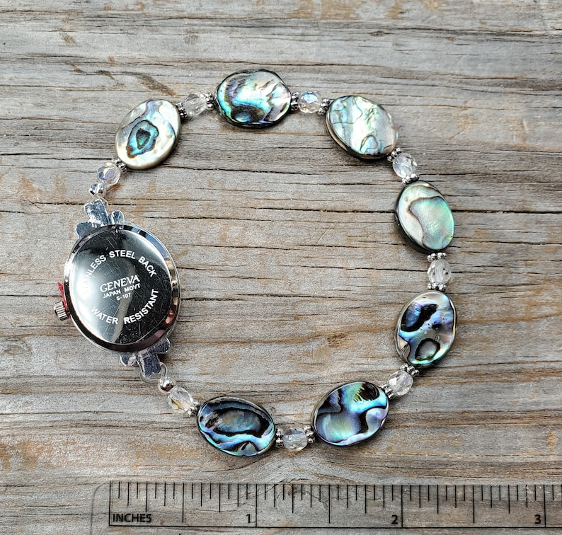 May include: A Geneva watch with a stainless steel back and water-resistant feature. The watch is attached to a bracelet made of iridescent abalone shells and clear beads. The bracelet is approximately 7 inches in length.