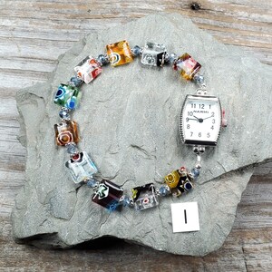May include: A wristwatch with a rectangular silver-tone case and a white face featuring the text NARMI and black numerals. The watch band consists of multi-colored square glass beads with floral patterns separated by small blue metallic spacers.