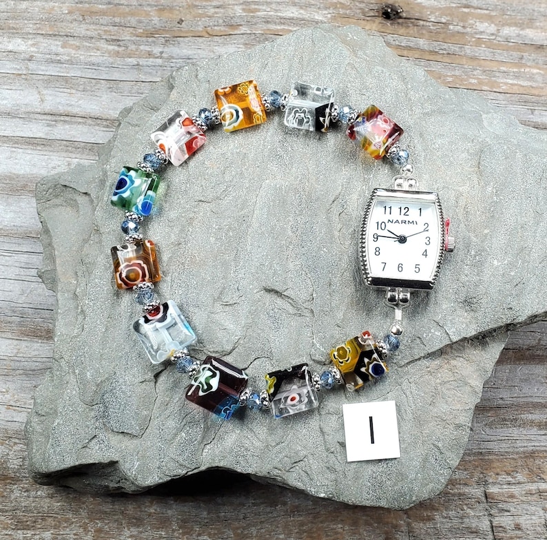 May include: A wristwatch with a rectangular silver-tone case and a white face featuring the text NARMI and black numerals. The watch band consists of multi-colored square glass beads with floral patterns separated by small blue metallic spacers.