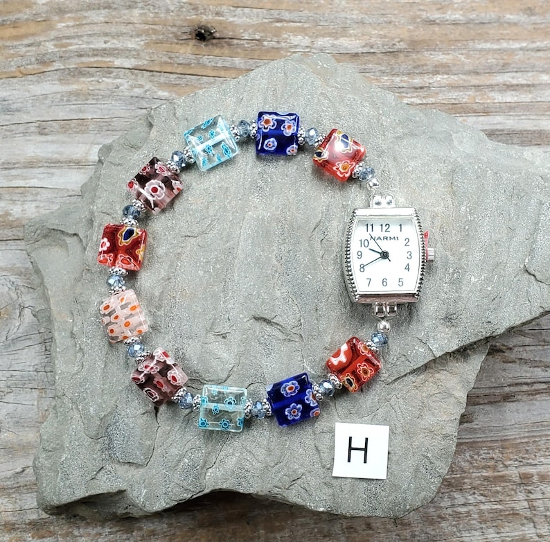 May include: A wristwatch with a rectangular silver-tone case and a white dial featuring the brand name Narmi. The watch band is composed of multi-colored square glass beads with floral patterns, separated by small silver-tone spacer beads.
