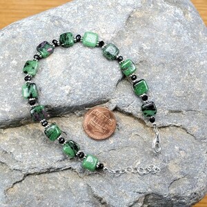 May include: A gemstone bracelet featuring square-cut green and red stones, interspersed with small black beads. The bracelet is displayed on a gray stone surface, with a penny for size reference. The bracelet has a silver-colored clasp.