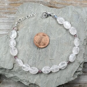 May include: A clear quartz bracelet with oval beads and small purple beads. The bracelet has a silver chain and clasp. A penny is next to the bracelet for size comparison.