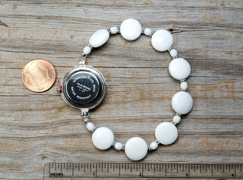 Puede incluir: Reloj de pulsera con brazalete de cuentas de cer&aacute;mica blanca y una esfera plateada que dice "NARMI JAPAN MVT L-37 WATER RESISTANT STAINLESS STEEL BACK".