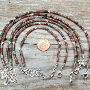 May include: A collection of beaded necklaces in shades of red, green, and brown. Each necklace is made with small, cylindrical beads and silver-coloured star-shaped embellishments. The necklaces are presented on a wooden surface.