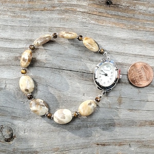 May include: A handmade watch bracelet featuring oval-shaped, cream-colored beads with brown marbling. The watch has a silver-toned case and a white face with black numerals. A penny is next to the watch for size comparison.