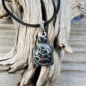 May include: A black turtle pendant necklace with iridescent accents. The pendant is made of glass and has a detailed carving of a turtle shell. The necklace is made of black cord.
