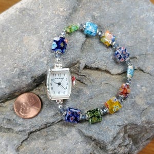 May include: A silver-toned watch with a white face and black numerals is attached to a bracelet of colorful glass beads. The beads are square and feature floral designs in various colors. A penny is next to the watch for scale.
