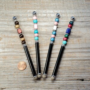 May include: Four black stylus pens with different colored beads. The beads are pink, blue, brown, and turquoise.