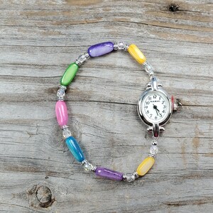 May include: A colorful beaded watch with a silver-toned watch face. The watch band features a variety of beads in shades of purple, green, pink, blue, and yellow. The watch face displays the time.