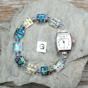 May include: A wristwatch with a rectangular silver-tone case and a white dial featuring black numerals and the text MARMI. The watch band consists of square glass beads with multi-colored floral patterns and small blue spacer beads. A letter G is visible.