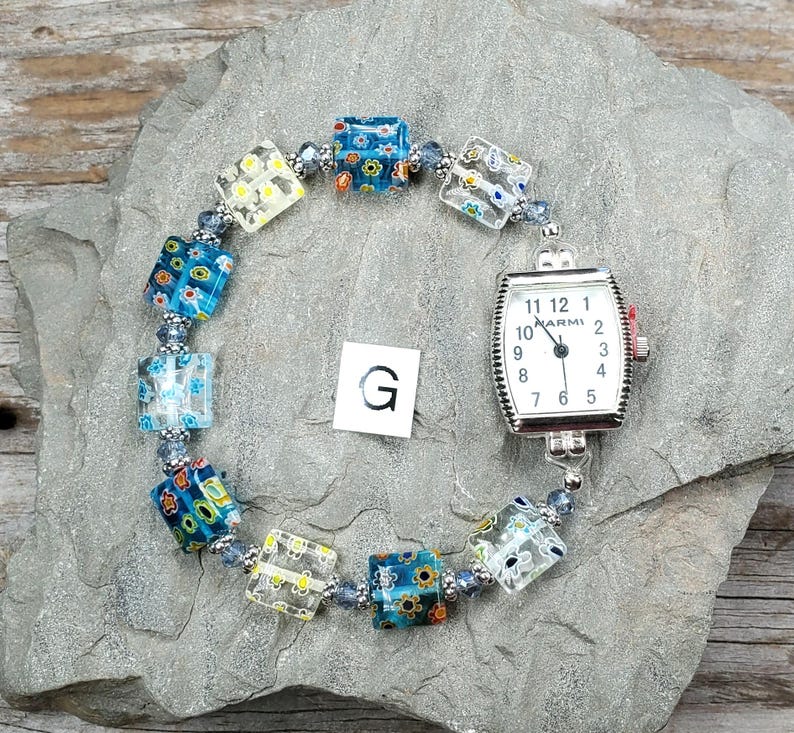 May include: A wristwatch with a rectangular silver-tone case and a white dial featuring black numerals and the text MARMI. The watch band consists of square glass beads with multi-colored floral patterns and small blue spacer beads. A letter G is visible.