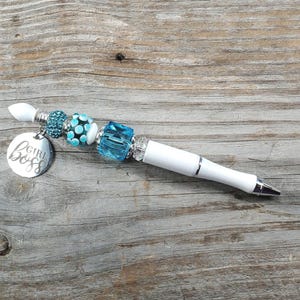 May include: A white pen with a silver tip and blue and clear decorative beads. A silver charm with the words "GIRL BOSS" hangs from the pen. The pen is set on a weathered wooden surface.
