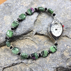 May include: A silver watch with a white face and black numbers. The watch is attached to a bracelet made of green and black square beads.